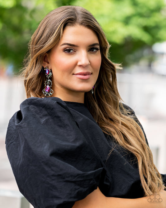 A cosmic collision of iridescent pink teardrop gems and a solitaire smoky emerald cut rhinestone haphazardly adorns studded silver frames. Dotted in round and emerald cut hematite rhinestone accents, the abstract frames stack into a stellar lure. Earring attaches to a standard post fitting. Due to its prismatic palette, color may vary. Sold as one pair of post earrings.