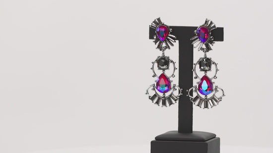 A cosmic collision of iridescent pink teardrop gems and a solitaire smoky emerald cut rhinestone haphazardly adorns studded silver frames. Dotted in round and emerald cut hematite rhinestone accents, the abstract frames stack into a stellar lure. Earring attaches to a standard post fitting. Due to its prismatic palette, color may vary.  Sold as one pair of post earrings.